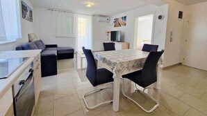 A modern, well-lit living room with a sofa, a dining table with chairs, a TV, and a kitchen area with a stove. - Holiday apartment newly furnished for 2-4 pers. with air conditioning, WLAN and car park (Baška)