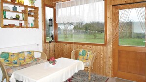 Ein gemütlicher Raum mit Holzvertäfelung, einem Tisch mit weißer Tischdecke, zwei Stühlen mit bunten Kissen, einem Regal mit Dekoration und einem großen Fenster mit Blick auf eine grüne Wiese.