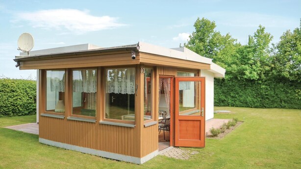 Ein kleines, eingeschossiges Holzhaus mit einem großen Fenster und einer Tür, umgeben von einem gepflegten Rasen und Bäumen.