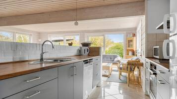 Eine moderne Küche mit Holzarbeitsplatte, Spüle aus Edelstahl und weißen Schränken. Es gibt einen Essbereich mit Tisch und Stühlen sowie einen Ausblick ins Freie durch große Fenster.