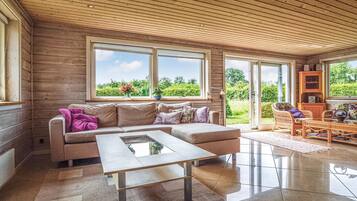 Ein modernes Wohnzimmer mit einer Couch, einem Couchtisch und Korbstühlen. Große Fenster bieten Blick auf Grünflächen.