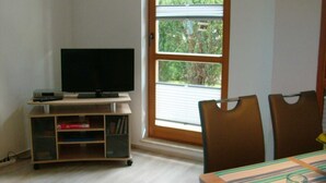 A room with a television on a wooden stand, a small bookshelf, and a dining table with chairs.