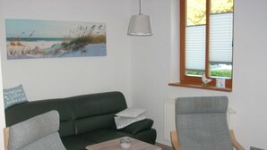 A living room with a green sofa, two grey armchairs, a small wooden table, a painting of a beach, a hanging lamp, and a window with blinds.