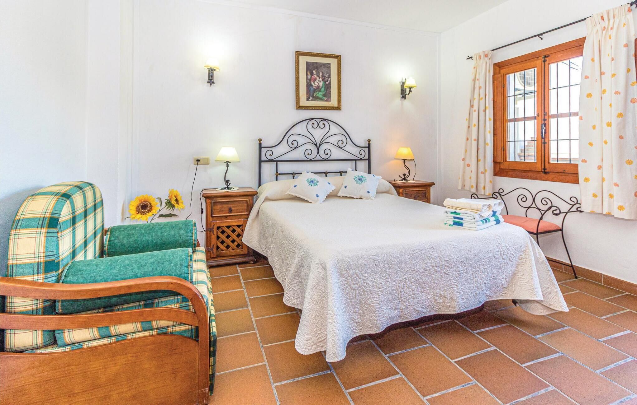 Une chambre à coucher avec un lit, une chaise, une table de chevet, une lampe, un tableau au mur et une fenêtre avec des rideaux.
