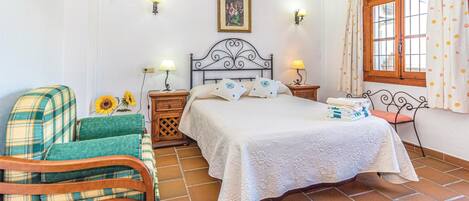 Une chambre à coucher avec un lit, une chaise, une table de chevet, une lampe, un tableau au mur et une fenêtre avec des rideaux.