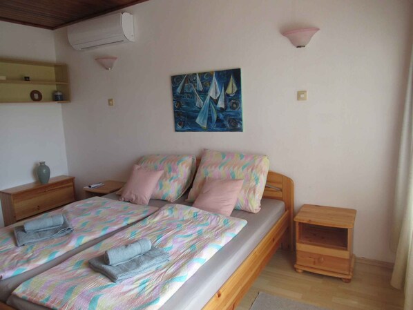 A bedroom with a bed, two bedside tables, a nightstand, a wall-mounted air conditioner, a shelf with decorative items, and a painting.