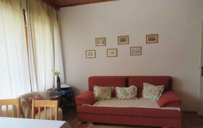 A living room with a red sofa, a wooden chair, a small table with a vase of flowers, and five framed pictures on the wall.