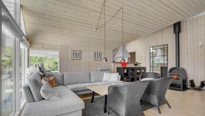 A modern living room with a grey sectional sofa, a dining table, wicker chairs, a fireplace, and wooden panel walls. - Nice home in Væggerløse with sauna (Væggerløse)