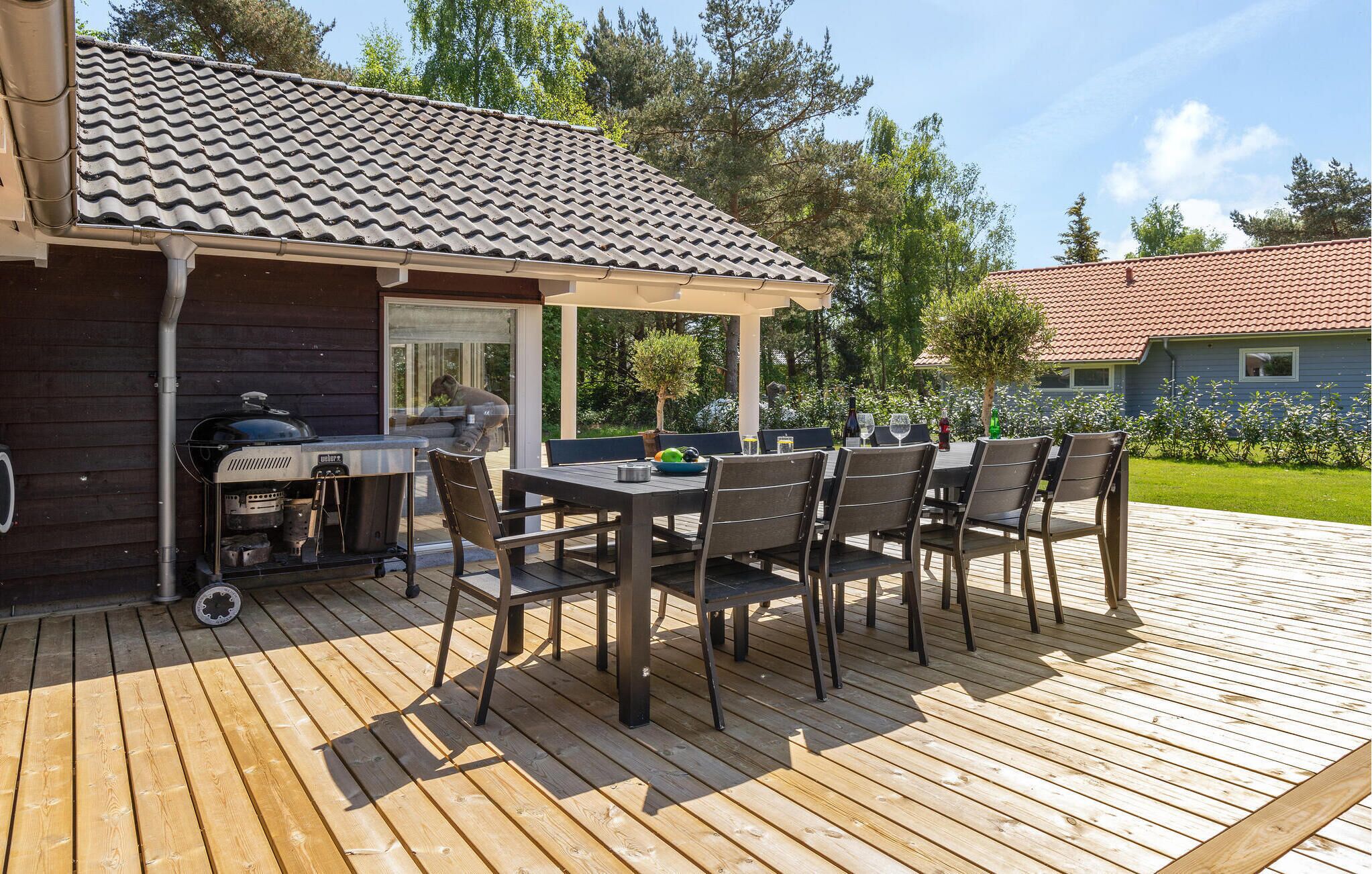 An outdoor deck with a grill and dining area, surrounded by trees and a house with a tiled roof.