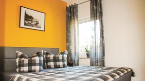 Ein Schlafzimmer mit einem Bett, karierten Bettbezügen, einem grauen Kopfteil, einem gerahmten Bild an der Wand und einem Fenster mit Vorhängen.