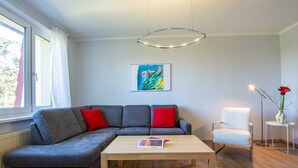 A modern living room with a grey sectional sofa, a wooden coffee table, a white armchair, and a floor lamp. A colorful abstract painting is on the wall, and a window with curtains is visible. - Dune garden Whg. Wa45-50 - Wa45-50 (Kühlungsborn)
