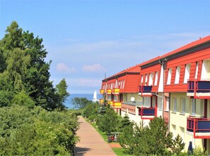 En rad röda och gula byggnader med balkonger, en asfalterad gångväg och en segelbåt i bakgrunden.