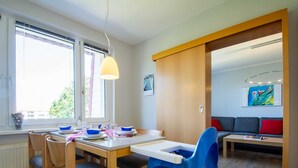 A dining area with a wooden table, blue chairs, and a blue high chair. There are windows with blinds, a sofa, and a painting on the wall. - Dune garden Whg. Wa45-50 - Wa45-50 (Kühlungsborn)