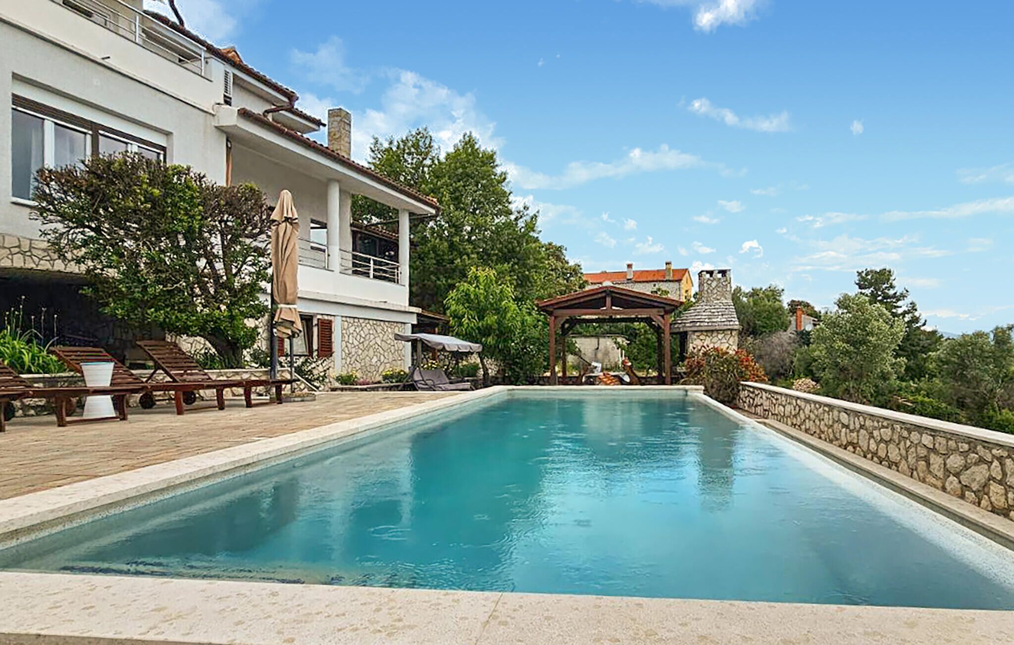 Une piscine dotée de chaises longues et d’une pergola, entourée de murs en pierre et de verdure.