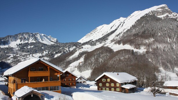 A mountain lodge with multiple wooden buildings, a snow-covered landscape, and a clear blue sky. - Vacation Apartment Lug - Gästehaus Margreth (Au)