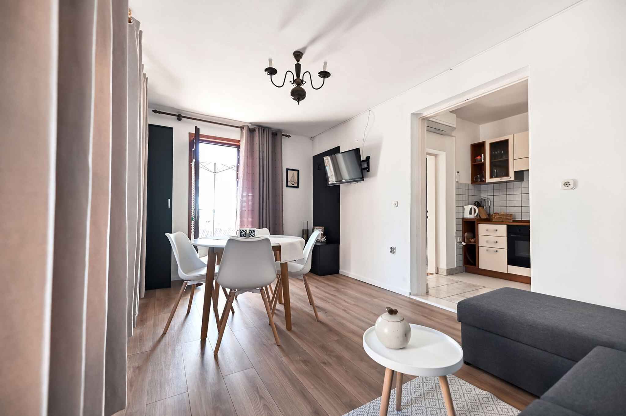 A modern living room with a grey sofa, a dining area with white chairs and a table, and a kitchen with cabinets and a TV.
