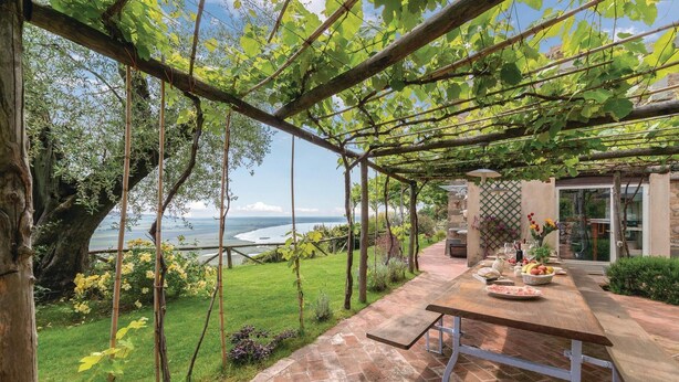 A covered outdoor dining area with a view of the sea, a stone wall, and a table set with food.