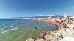 A crowded beach with people swimming and sunbathing, a Ferris wheel, and mountains in the background.
