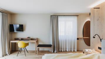 A modern hotel room with a wooden desk, a yellow chair, a bed with a white blanket, a TV, and a large mirror.