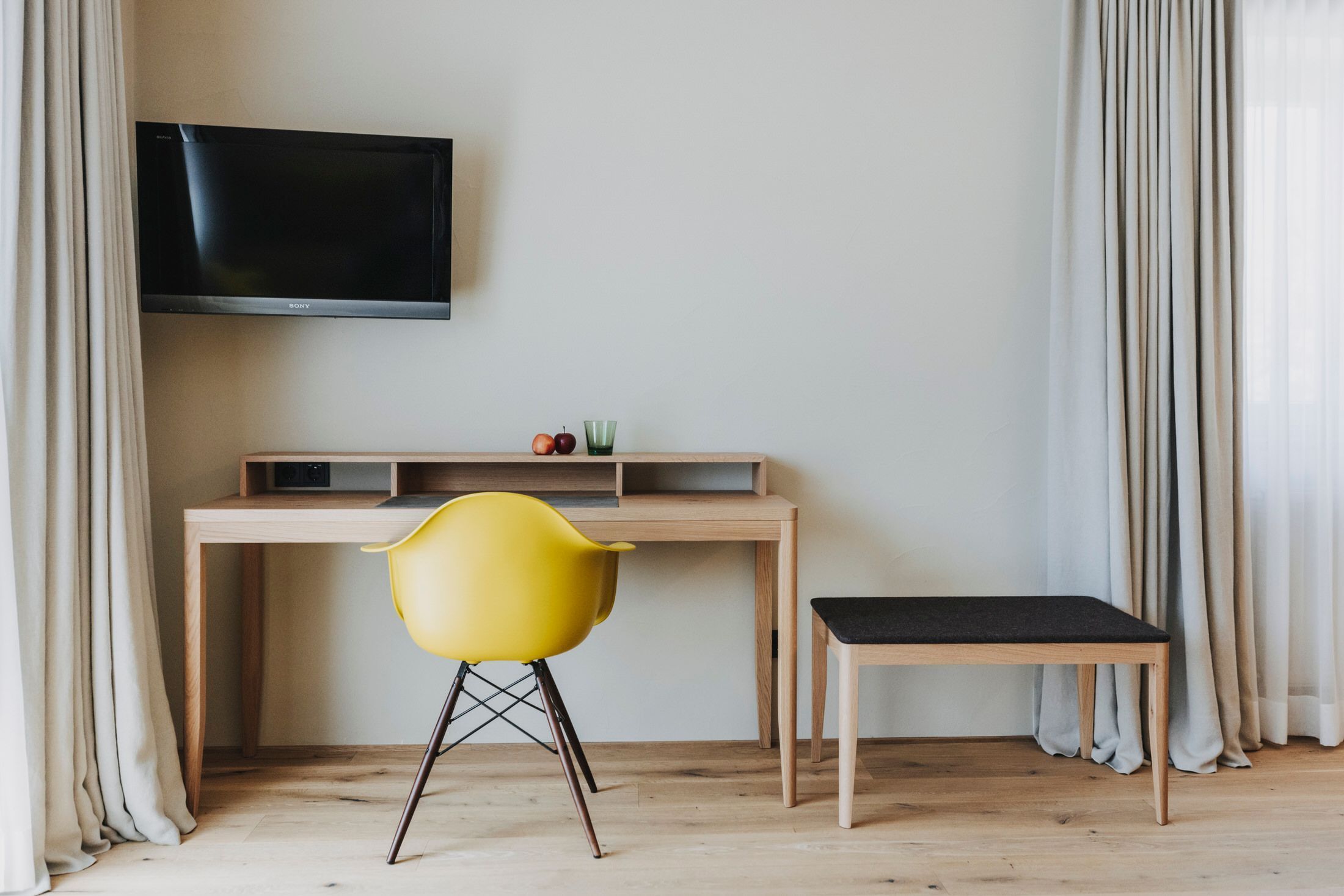 A room with a wooden desk, a yellow chair, a black bench, and a mounted TV.