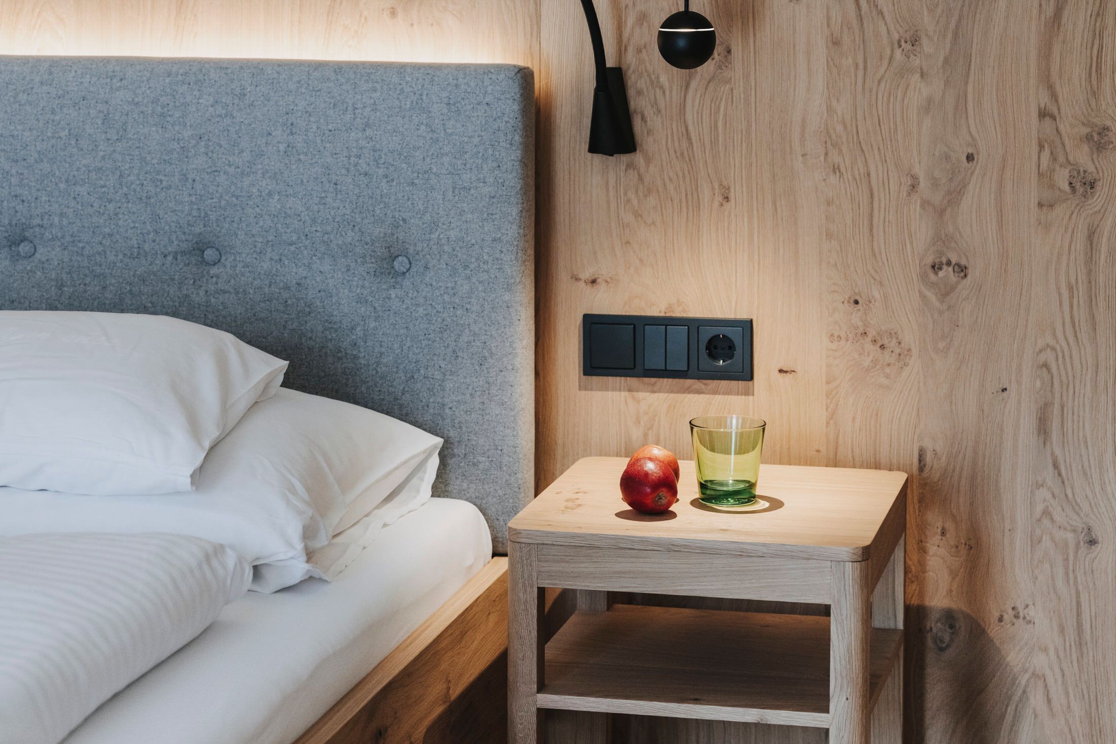 Una cama bien hecha con ropa blanca, una mesita de noche con enchufe, una lámpara y fruta con un vaso sobre la mesa.