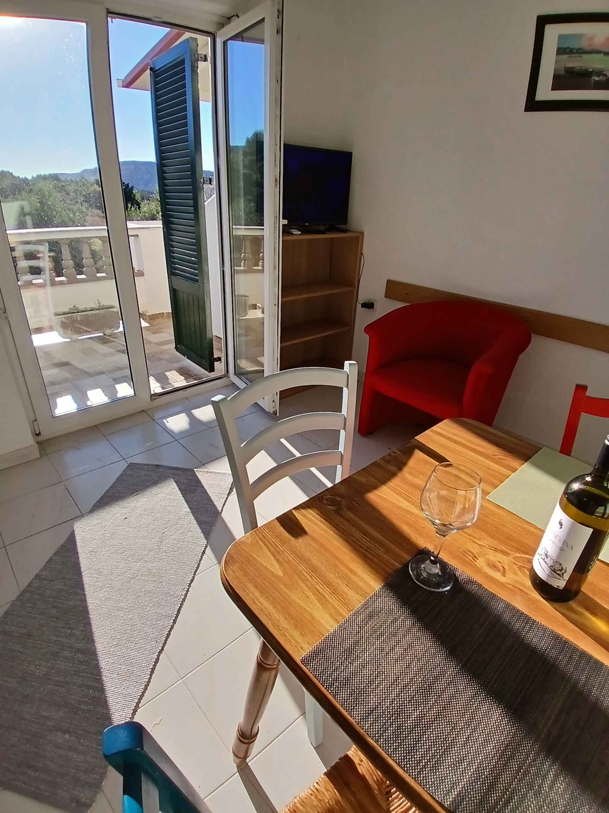 Una habitación con una mesa de comedor de madera, una silla roja, un televisor sobre un mueble bajo y vistas a un área exterior con balcón a través de una puerta de cristal.