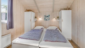 Um pequeno quarto estilo cabana, com duas camas de solteiro, uma janela com cortinas e uma prateleira na parede com uma planta.