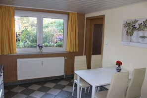 Une pièce avec une fenêtre, un radiateur, une table avec des chaises et un vase rempli de fleurs.