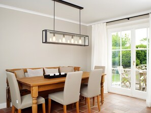 Un comedor con mesa y sillas de madera, una lámpara colgante y vista a un jardín a través de grandes puertas de vidrio.