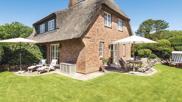 Une maison en briques avec un toit de chaume, une terrasse avec des meubles d’extérieur et une pelouse bien entretenue.