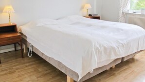 Cama bem arrumada em quarto com piso de madeira, duas mesas de cabeceira com luminárias, um quadro na parede e uma janela com cortinas.