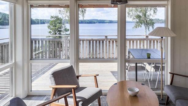 A room with large windows overlooking a lake, a wooden deck, and a table with chairs.