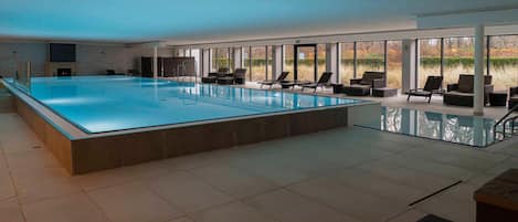 A large indoor swimming pool with clear glass sides, surrounded by lounge chairs and sofas, and large windows offering views of the outdoors.