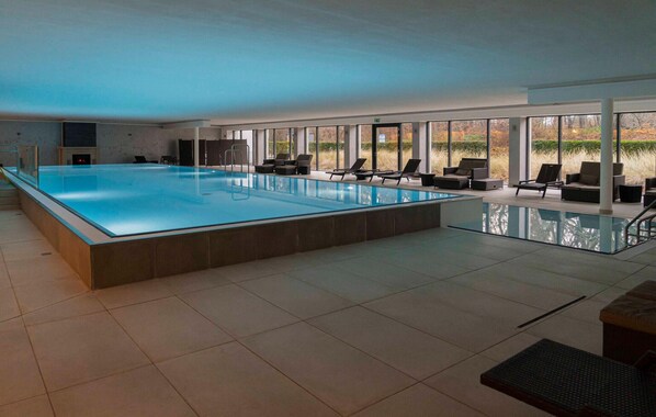 Ein großer, überdachter Pool mit klaren Glaswänden, umgeben von Liegestühlen und Sofas, mit großen Fenstern, die einen Blick ins Freie bieten.