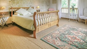 Un dormitorio con cama de madera, pared de piedra, ventana con un trípode y alfombra estampada.