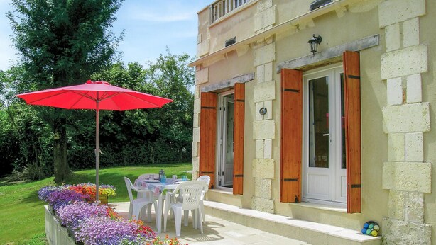 Un patio con una sombrilla roja, sillas blancas y una mesa. Hay macizos de flores con diversas flores.
