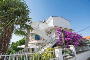 Uma casa branca de dois andares com escadaria de corrimão branco, varanda e uma árvore com flores roxas.