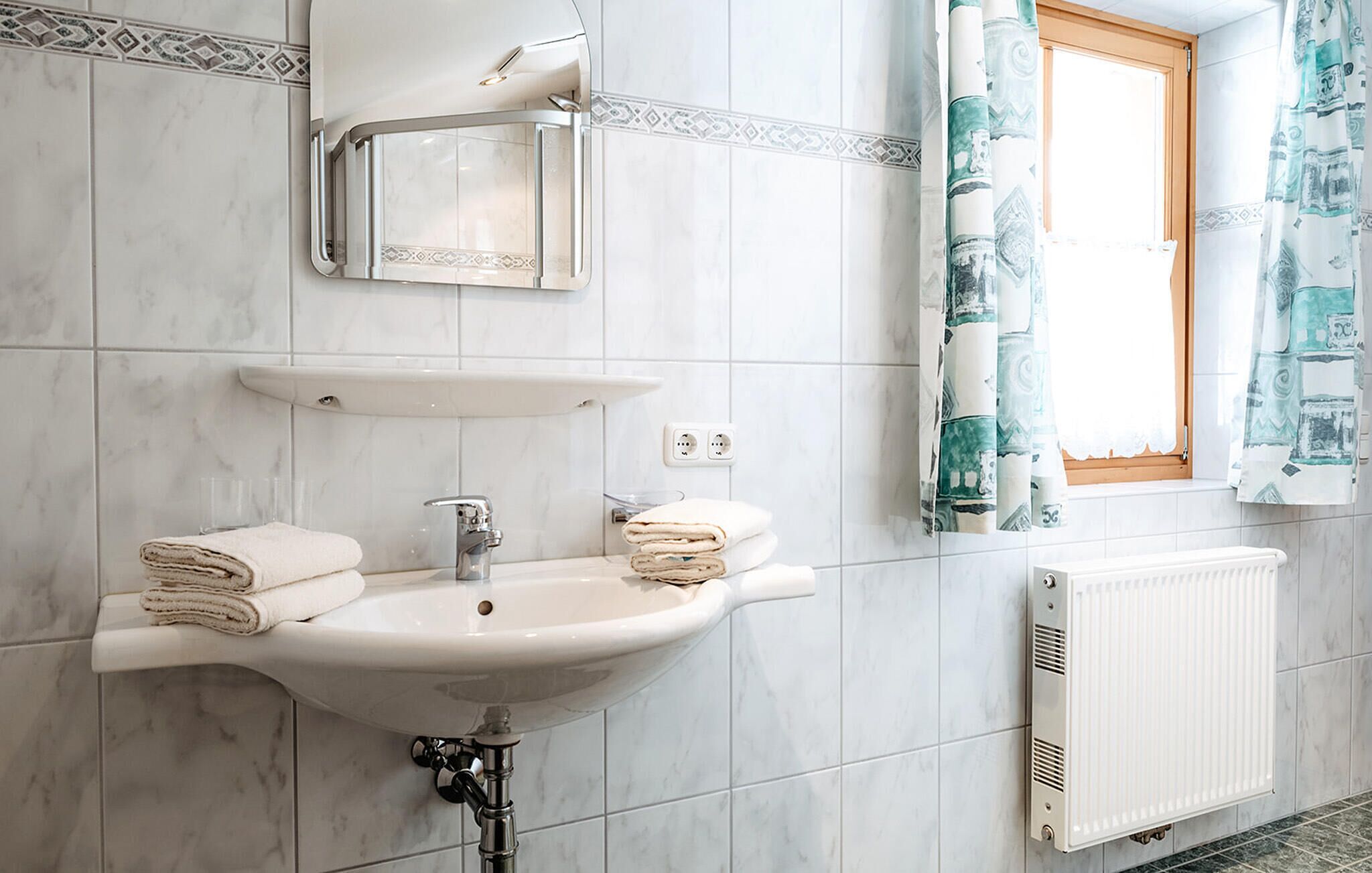 Un baño con lavamanos blanco, espejo y toallero. Se ve una ventana con cortinas y un radiador en la pared.