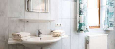 Un baño con lavamanos blanco, espejo y toallero. Se ve una ventana con cortinas y un radiador en la pared.