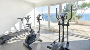 A home gym with cardio equipment including a treadmill, elliptical, and stationary bike, overlooking a scenic view of the sea. - 1 bedroom nice apartment in Stanici (Stanici)