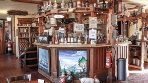Una zona de bar con estantes de madera llenos de botellas y vasos, una chimenea con un cuadro, y un extintor.