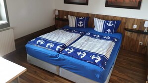 Cama doble con ropa de cama azul y diseños de anclas, cabecera de madera, dos cuadros colgados en la pared y una mesita de noche con lámpara.