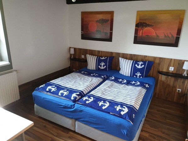 Cama doble con ropa de cama azul y diseños de anclas, cabecera de madera, dos cuadros colgados en la pared y una mesita de noche con lámpara.