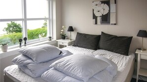 A neatly made bed with white bedding and multiple pillows, a bedside table with a lamp, a small potted plant, and a window with a view of greenery. - Awesome home in Rudkøbing (Rudkøbing)