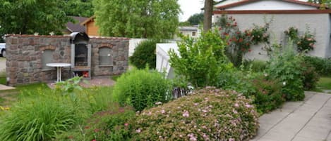 A stone barbecue area with a table and chairs, surrounded by a garden with various plants and flowers.