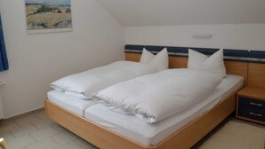 Una cama individual con ropa blanca, cabecera con una franja azul, mesita de noche con cajón y un cuadro en la pared.