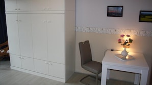 Een kamer met een grote witte garderobe, een klein wit tafeltje met een bloemenvaas, een grijze stoel en twee foto's in lijsten aan de muur.
