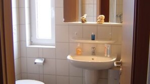 Een kleine badkamer met toilet, wastafel en spiegel. Boven het toilet bevindt zich een raam en er is een klein plankje met toiletartikelen.