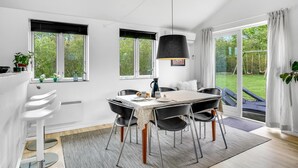 Et moderne spiseområde med et bord dækket til fire personer, omgivet af sorte stole, en hængende sort lampe og udsigt til en have gennem store vinduer.