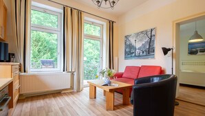 Ein modernes Wohnzimmer mit einem roten Sofa, einem schwarzen Sessel und einem Holztisch. Es gibt ein großes Fenster mit Vorhängen, eine Heizung und eine Pendelleuchte.